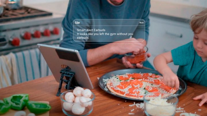 Tile ad scene: a Smart Alert notification that earbuds were left at the gym, shown on a tablet in a family kitchen